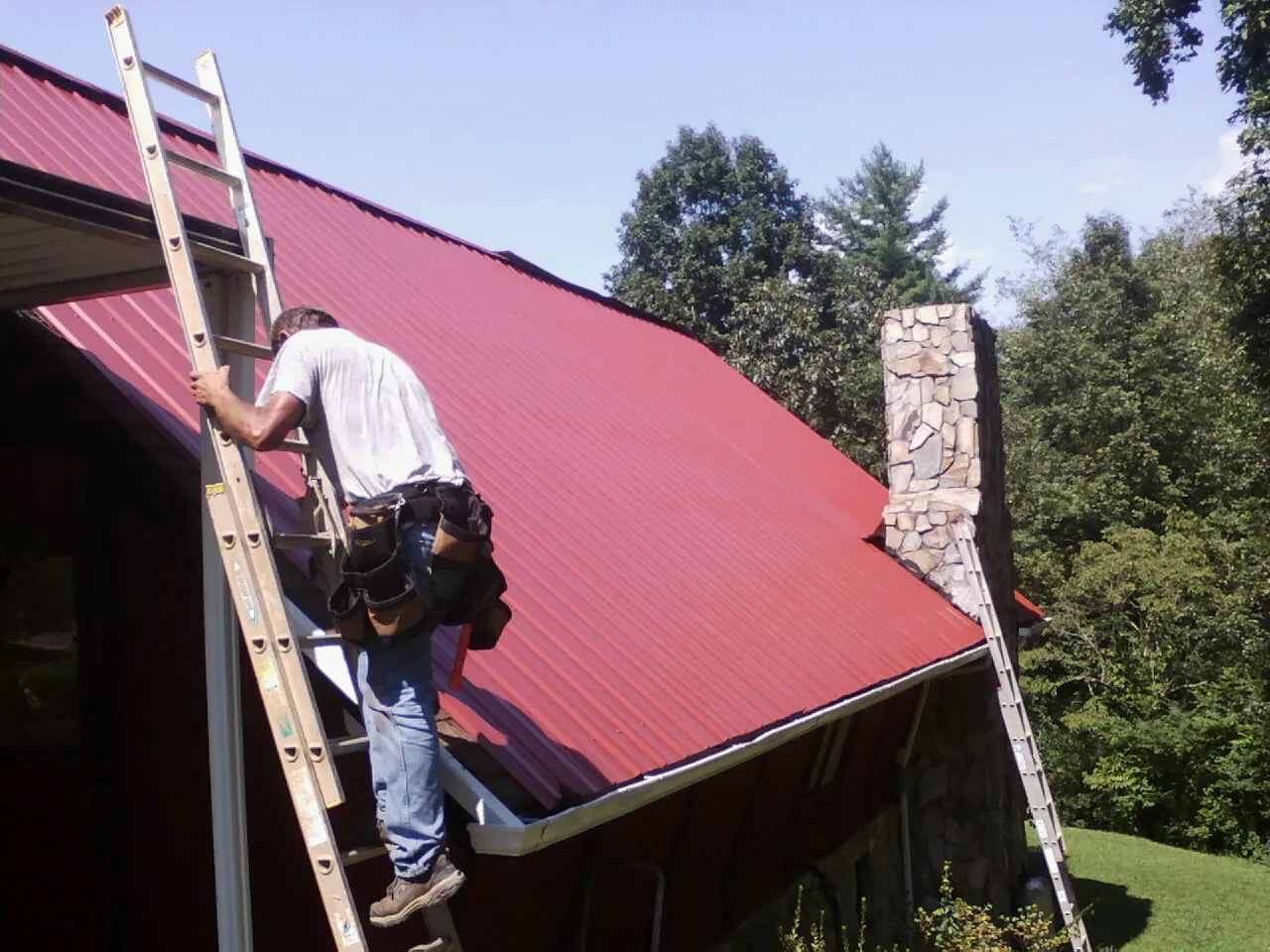 Roofer on a ladder beside a red metal roof and stone chimney — Soffit & Fascia Repair by Roofing Brothers in Baconton